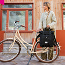 Urbane Radfahrerin hält ein schwarzes WEATHERGOODS SWEDEN Bikepack am Tragegriff neben ihrem Citybike vor einer modernen Holzfassade.